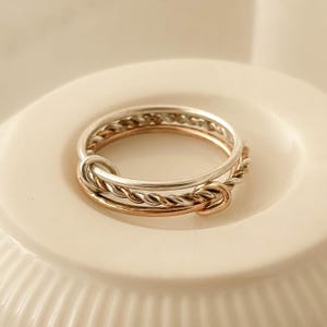 Puede incluir: Un conjunto de anillos apilables con tres anillos en tonos plateados y dorados. Los anillos tienen un diseño de cuerda trenzada y un detalle de nudo, creando un efecto en capas. Los anillos se muestran sobre una superficie blanca.