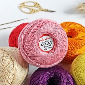 May include: Six balls of embroidery floss in various colors, including pink, yellow, orange, red, cream, and purple. The pink ball has a label that reads "col. 0341 MAJA 5 120 tex x 2 50g/210m".