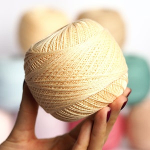 以下が含まれることがあります： 淡いベージュ色の糸玉を手に持っています。背景には、さまざまなパステルカラーの糸玉がぼやけています。編み物や編み物プロジェクトに適した、綿または同様の繊維のようです。