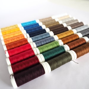 Puede incluir: Una colección de carretes de hilo de varios colores, incluyendo rojo, verde, azul, amarillo y marrón. Los carretes están dispuestos en un patrón escalonado, con los extremos del hilo visibles.