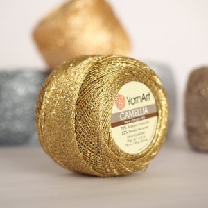 Könnte beinhalten: Ein Garnball in Gold und Silber mit einem Etikett mit der Aufschrift "YarnArt Camellia 70% Polyester, 30% Metallic, www.yarnart.info, 20 g, 100 m (109 yds)"