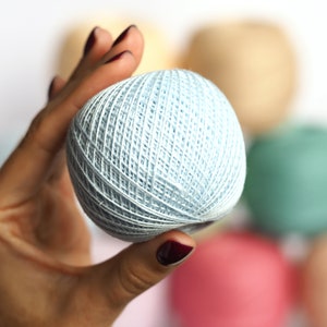 Puede incluir: Una mano sosteniendo una bola de hilo azul claro. El hilo est&aacute; enrollado en forma de bola y tiene una superficie texturizada. La bola de hilo est&aacute; enfocada, mientras que el fondo est&aacute; desenfocado.