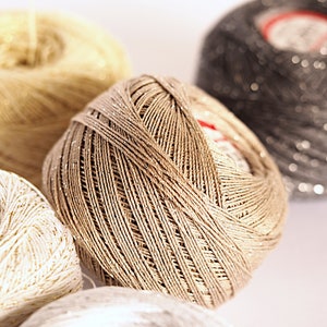 May include: Four spools of yarn in different colours and textures. The spools are beige, white, and grey with a silver shimmer.