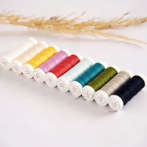 Peut inclure: Une collection de 10 bobines de fil de différentes couleurs, dont blanc, crème, jaune, rose, rouge, bleu clair, sarcelle, vert, beige et noir. Les bobines sont disposées en rangée de gauche à droite, la bobine blanche à gauche et la bobine noire à droite.