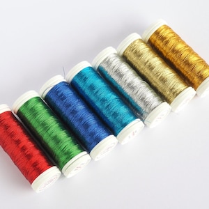 Può includere: Un set di sette bobine di filo metallico in vari colori, tra cui rosso, verde, blu, blu turchese, argento, oro e un oro chiaro. Le bobine sono disposte in fila da sinistra a destra, con la bobina rossa a sinistra e la bobina oro chiaro a destra.