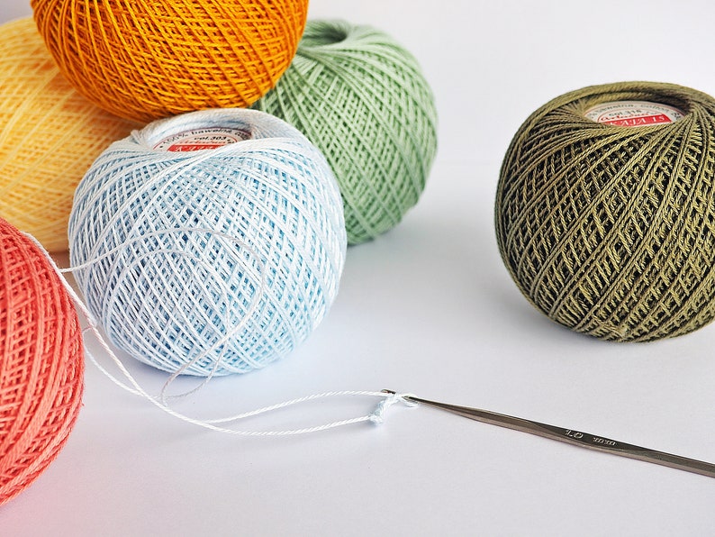 Puede incluir: Una colecci&oacute;n de ovillos de lana de colores en naranja, amarillo, verde, azul y rojo. Un ganchillo blanco est&aacute; sobre una superficie blanca.