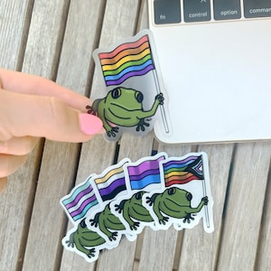 May include: A set of six frog stickers with different pride flags. The frogs are holding the flags in their hands. The flags include the rainbow pride flag, the transgender pride flag, the bisexual pride flag, the pansexual pride flag, the genderfluid pride flag, and the asexual pride flag.