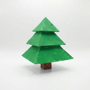 May include: A handmade, green Christmas tree decoration. The tree is constructed from three stacked, triangular layers, painted in a vibrant green hue. It is mounted on a small, square, brown wooden base. The tree is a festive, handcrafted item.