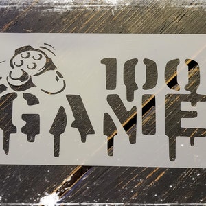 Könnte beinhalten: Eine weiße Schablone mit einem schwarzen Design eines Videospiel-Controllers und dem Text "100% Gamer" mit tropfenden Farbeffekten.