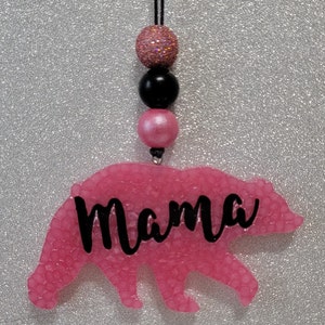 May include: Pink bear-shaped car charm with the word "Mama" written in black. The charm is attached to a black string with pink and black beads.