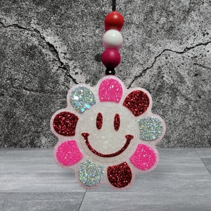 May include: A flower-shaped car charm with a smiling face design. The charm features a white center with a glittery outline and petals in pink, red, and silver glitter. It hangs from a black cord with red, white, and pink beads.