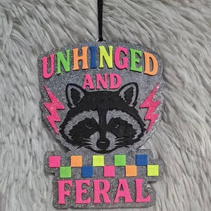 May include: A glittery, raccoon-themed ornament with the words "UNHINGED AND FERAL" in neon colors. The raccoon is black and white, with pink lightning bolts on either side. The ornament is suspended by a black string.