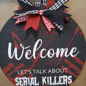 May include: A black round door hanger with red blood splatter and the text "Welcome Let's Talk About Serial Killers" in white. A black and white checkered bow with red ribbon accents is attached to the top.