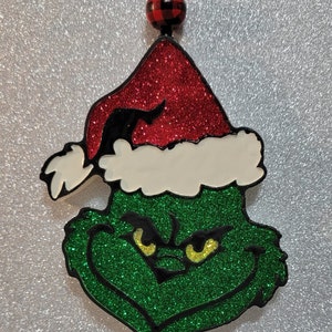 May include: Green Grinch Christmas ornament with a red Santa hat and black trim. The ornament is covered in glitter and has yellow eyes.