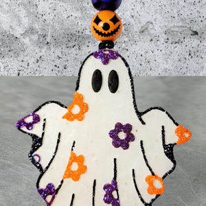 May include: A white ghost ornament with orange and purple glitter flowers. The ghost has black eyes and is attached to a string with black and white spiderweb, blue plaid, and orange pumpkin beads.