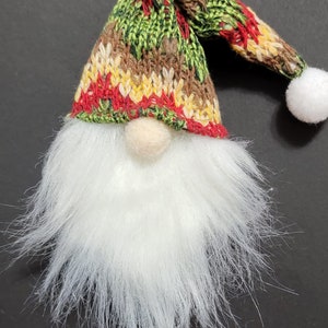 May include: A small gnome ornament with a white fuzzy beard and a knitted hat with red, green, brown, and yellow stripes. The hat has a white pom-pom on top.