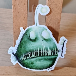 Puede incluir: Una pegatina con una ilustración de acuarela de un rape verde con un señuelo brillante. El pez tiene ojos grandes y dientes afilados. La obra de arte es de estilo acuarela con un cuerpo verde oscuro. Firma del artista.