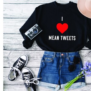 May include: Black sweatshirt with the text "I ❤️ MEAN TWEETS" in white and a red heart. The image also includes denim shorts, black and white trainers, a vintage camera, sunglasses, and a beige hat, suggesting a casual outfit.