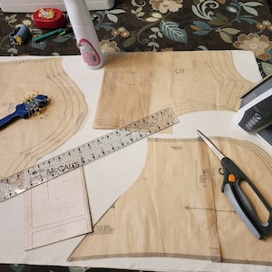 May include: A sewing project in progress with fabric patterns, a ruler, scissors, an iron, and a pin cushion.