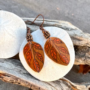 May include: A pair of copper leaf earrings with a brown and orange leaf design. The earrings are hanging from a copper wire with a small copper bead at the top.