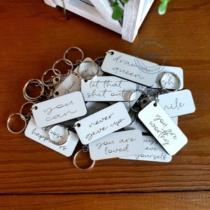 Könnte beinhalten: Ein Satz weißer Schlüsselanhänger aus Metall mit inspirierenden Botschaften, darunter "you can", "let that shit out", "never give up", "you are loved", "you are worthy", "draw queen", "choose happiness" und "smile".