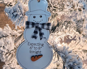 Op de afbeelding: Witte sneeuwman-versiering met een zwart-wit geruite sjaal en een wortelneus. De versiering heeft de tekst "Expecting 6" to 8" tonight".