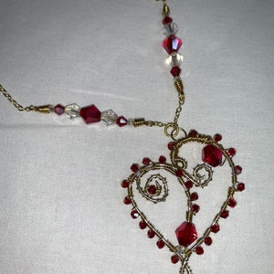 Puede incluir: Un collar de alambre dorado con un colgante en forma de corazón. El colgante está adornado con cuentas de cristal rojas y transparentes. La cadena y el alambre del collar son dorados, creando un accesorio romántico y elegante.