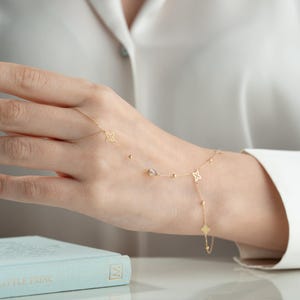 Può includere: Un braccialetto a catena d'oro con un delicato design a stella e un piccolo ciondolo di pietra preziosa. Il braccialetto viene indossato al polso e si estende fino al dito.