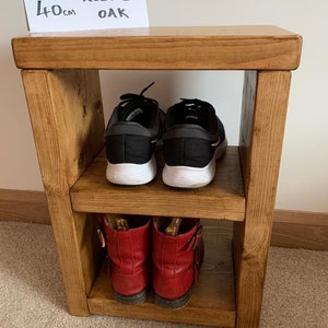 May include: A two-tier wooden shoe rack made of rustic oak. The rack is 40 cm tall and has two shelves for storing shoes. The top shelf has a pair of black trainers, and the bottom shelf has a pair of red boots.