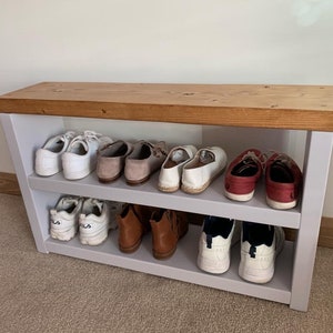 May include: A white shoe rack with two shelves and a wooden top. The rack is filled with various shoes, including white trainers, brown boots, and red sandals.