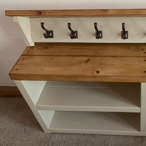 May include: White wooden hallway bench with a top shelf and four hooks. The bench has two shelves for storing shoes. The top shelf is made of wood and has a natural finish. The bench is painted white.