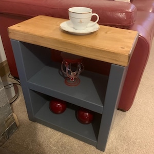 May include: A small wooden side table with a dark grey finish. The table has two shelves and is decorated with a white teacup and saucer, a red glass vase, and two red decorative balls.