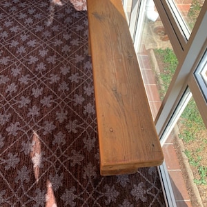 May include: A rectangular wooden bench with a weathered appearance, showing wood grain and nail heads. The bench is light brown and appears to be made from reclaimed wood. It is positioned next to a window.
