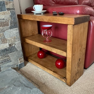 May include: A wooden side table with two shelves. The table is stained a light brown colour and has a rustic look. The table is next to a red leather sofa and has a red glass bowl and two red ornaments on the shelves.