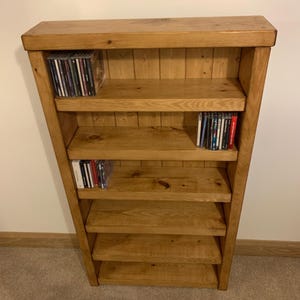 May include: A wooden bookcase with five shelves, displaying several CDs. The bookcase is constructed from light-coloured wood and features a subtly angled design. The shelves are evenly spaced, and the unit rests on a carpeted floor.