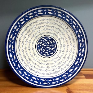 Op de afbeelding: Een ronde keramische serveerschaal met een blauw en wit visontwerp. De schaal heeft concentrische cirkels van zwemmende vissen, met een centrale cluster van vissen. De rand is donkerblauw. De schaal staat op een houten oppervlak.