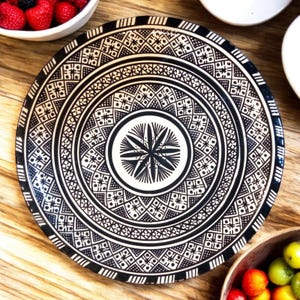 May include: A large, round ceramic plate with a black and white geometric pattern. The plate has a central design of a stylized flower with eight petals.