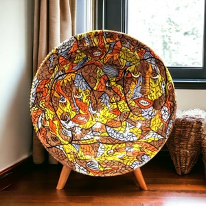 May include: A large, round ceramic plate with a colorful, abstract design. The plate features a mosaic of faces, birds, and geometric shapes in shades of yellow, orange, brown, and blue. The plate is on a wooden stand.