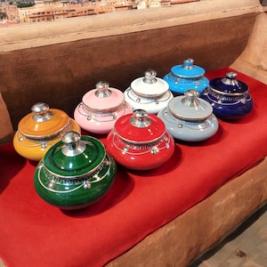 Puede incluir: Ocho botes de cerámica decorativos con tapa en varios colores, incluyendo verde, rojo, azul, blanco, amarillo y rosa. Cada bote tiene una tapa plateada con un borde decorativo.