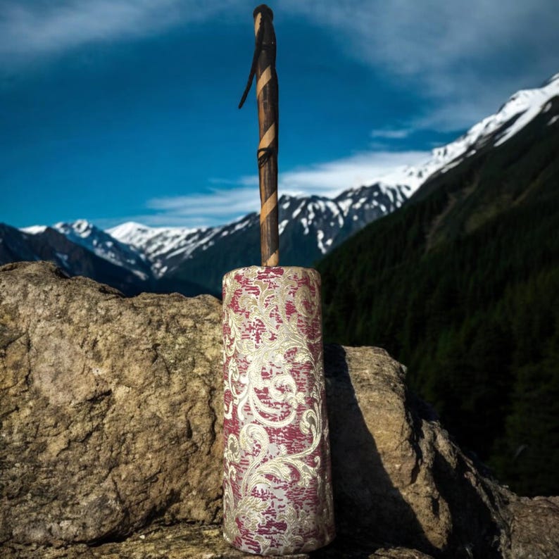 May include: A red and gold patterned fabric covered cylinder with a wooden stick attached to the top. The cylinder is sitting on a large rock with a mountain range in the background.