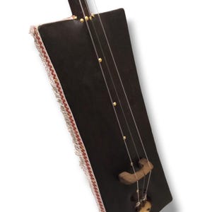 K&ouml;nnte beinhalten: Ein dunkelbraunes Holzmusikinstrument mit einem rechteckigen Korpus und einem langen Hals. Der Korpus ist mit einem wei&szlig;en und roten Fransenbesatz verziert. Das Instrument hat einen Steg aus Holz und ein rundes Schallloch.