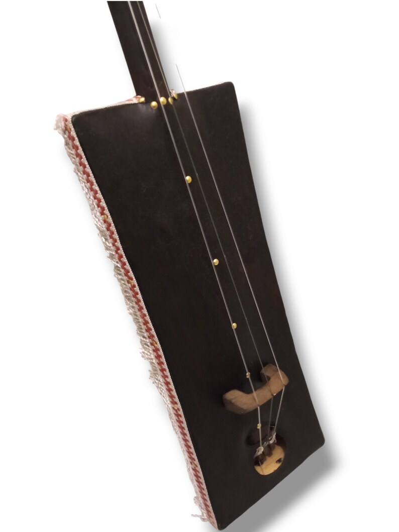 K&ouml;nnte beinhalten: Ein dunkelbraunes Holzmusikinstrument mit einem rechteckigen Korpus und einem langen Hals. Der Korpus ist mit einem wei&szlig;en und roten Fransenbesatz verziert. Das Instrument hat einen Steg aus Holz und ein rundes Schallloch.