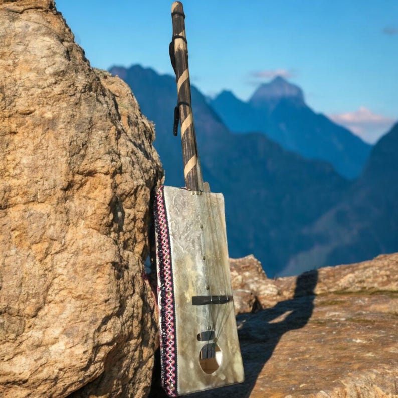 May include: A traditional stringed instrument with a wooden neck and a rectangular body made of animal skin. The instrument is decorated with a colorful patterned trim and is leaning against a large rock. The background is a blurry image of mountains.