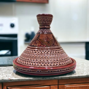 May include: A brown and red ceramic tagine with intricate designs and a conical lid. The tagine is a traditional Moroccan cooking pot.