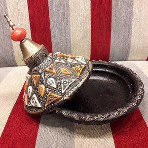 Puede incluir: Un recipiente decorativo negro y plateado abierto con tapa. La tapa está adornada con formas geométricas coloridas y un remate rojo y dorado. El recipiente se encuentra sobre una superficie a rayas rojas y grises.