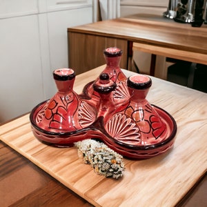 May include: A red ceramic tagine with a unique three-section design. The tagine features intricate black and white floral patterns and a dark brown rim. The lids have a conical shape. The tagine is displayed on a wooden cutting board.
