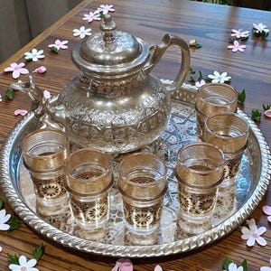 Könnte beinhalten: Ein silbernes Teeservice mit einer dekorativen Teekanne und sechs kleinen Gläsern, alles auf einem silbernen Tablett. Die Gläser haben goldene Ränder und florale Muster. Das Set ist auf einem Holztisch mit verstreuten Blumendekorationen angeordnet.