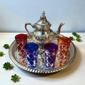 Könnte beinhalten: Ein silbernes Teeservice mit einer dekorativen Teekanne und einem runden Tablett mit sechs bunten Gläsern. Die Gläser sind rot, blau, lila und orange, mit einem silbernen Filigranmuster. Das Set steht auf einer weißen Oberfläche.