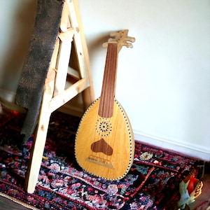 May include: A wooden oud, a type of lute, with a light brown body and a dark brown neck. The oud is resting on a colourful rug with a wooden stand behind it.