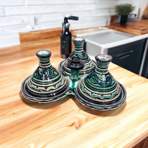 Op de afbeelding: Drie groene en blauwe keramische tagines met ingewikkelde ontwerpen staan op een houten oppervlak. De tagines zijn in een cirkelvormige opstelling geplaatst met een centrale knop.